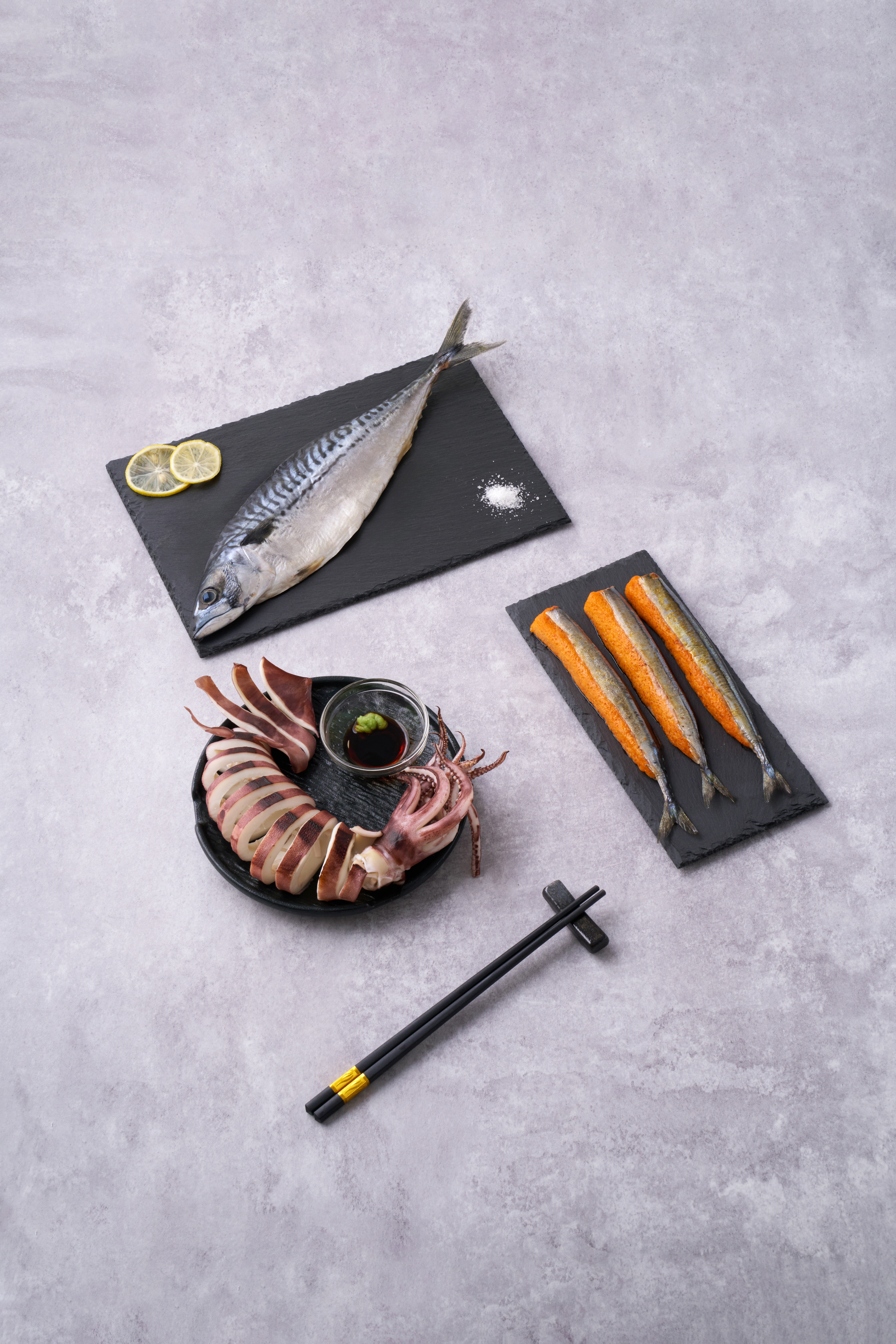 Mackerel and saury — overhead studio shot showing a whole Pacific mackerel on a black slate, sliced bullet squid composition on a black plate, and three saury fillets topped with mentaiko on a slate board.