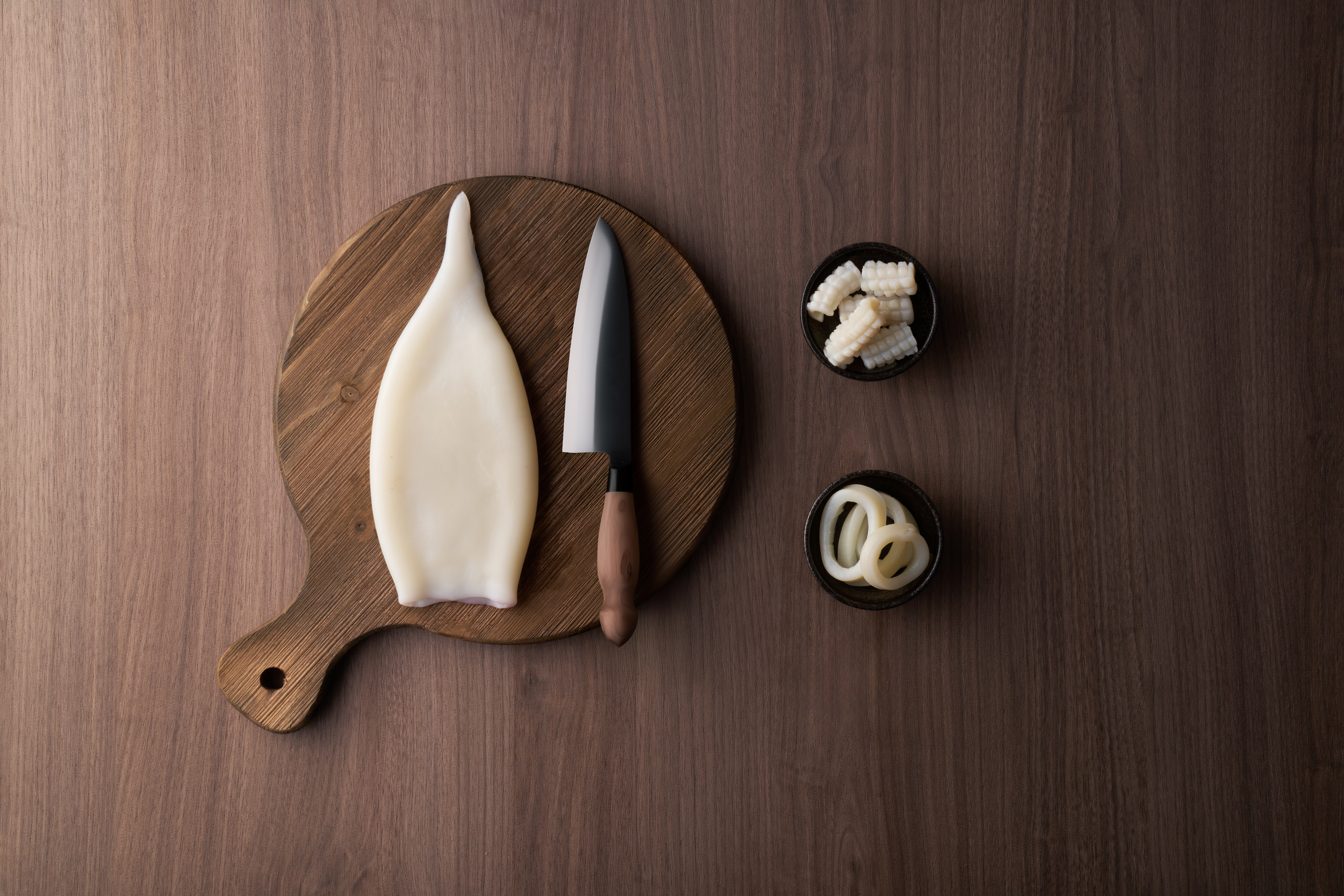 Secondary cutting — overhead studio shot of a cleaned squid tube on a round wooden cutting board with a chef's knife, alongside small dishes of diced squid and squid rings.