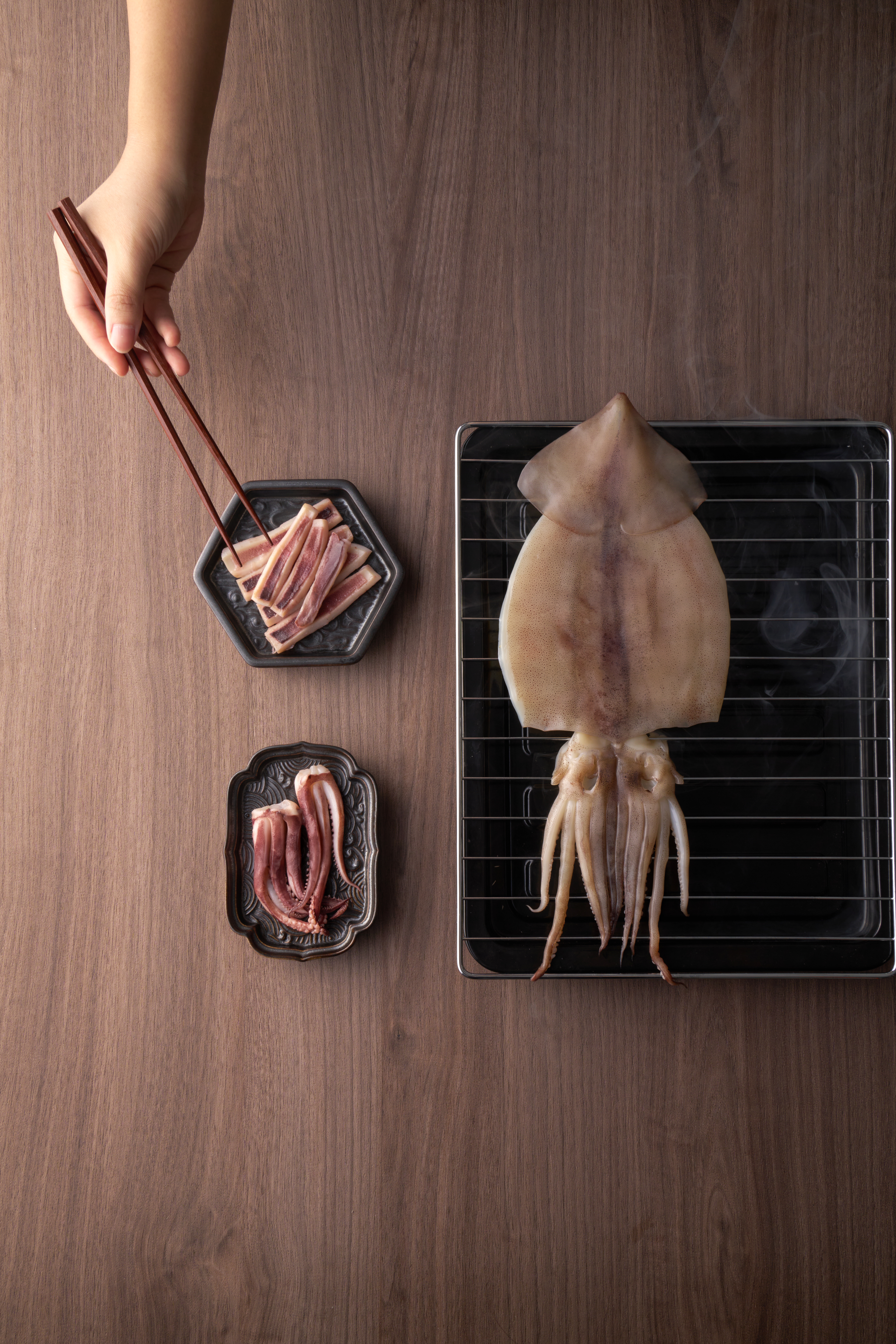 Seasoning tier — overhead studio shot of a marinated cleaned squid on a metal grilling rack with chopsticks lifting strips of marinated squid from a small black dish.